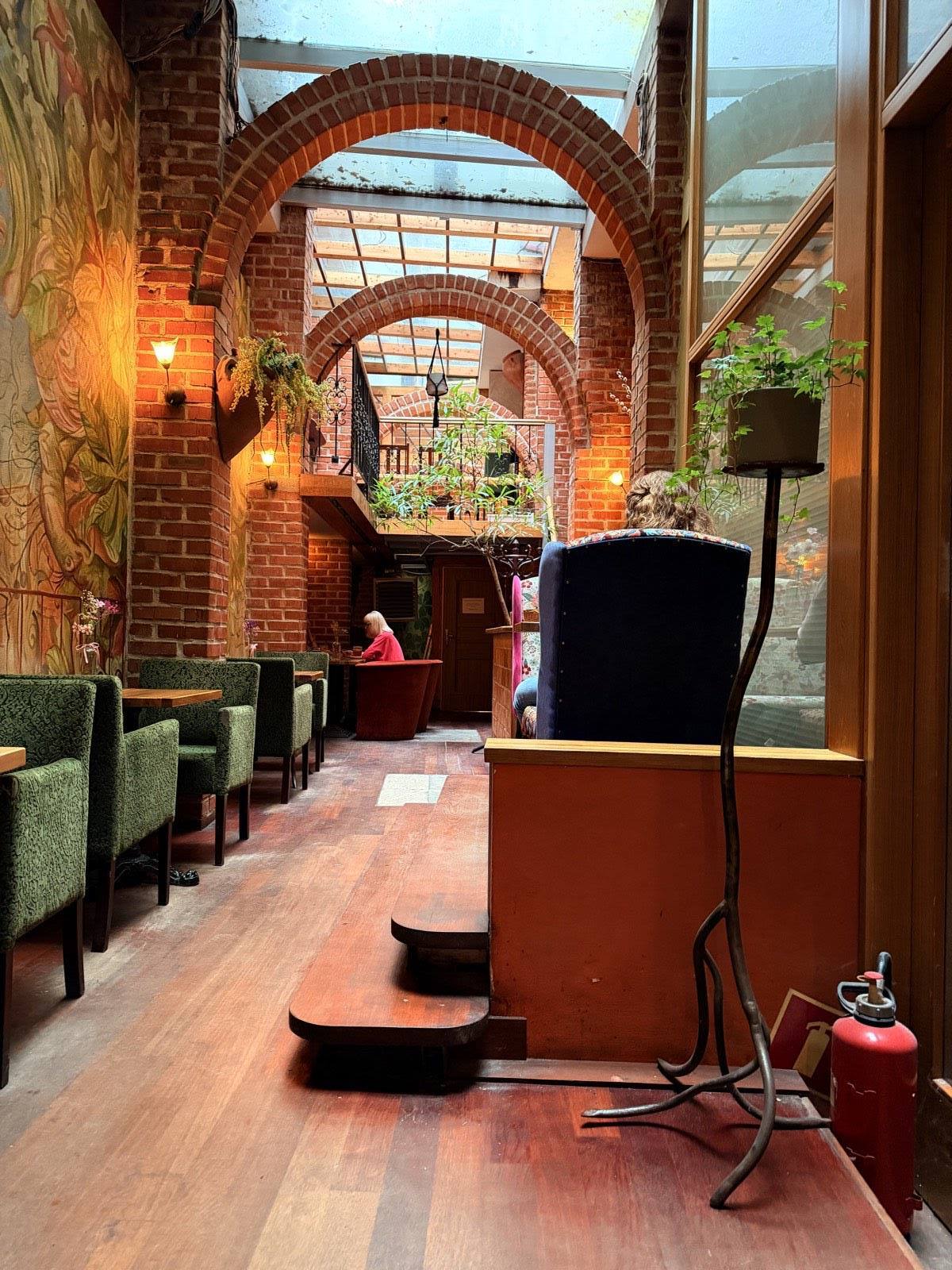 The garden atrium — red-brick arches rising to a glass skylight roof, green velvet armchairs and a painted mural along the wall